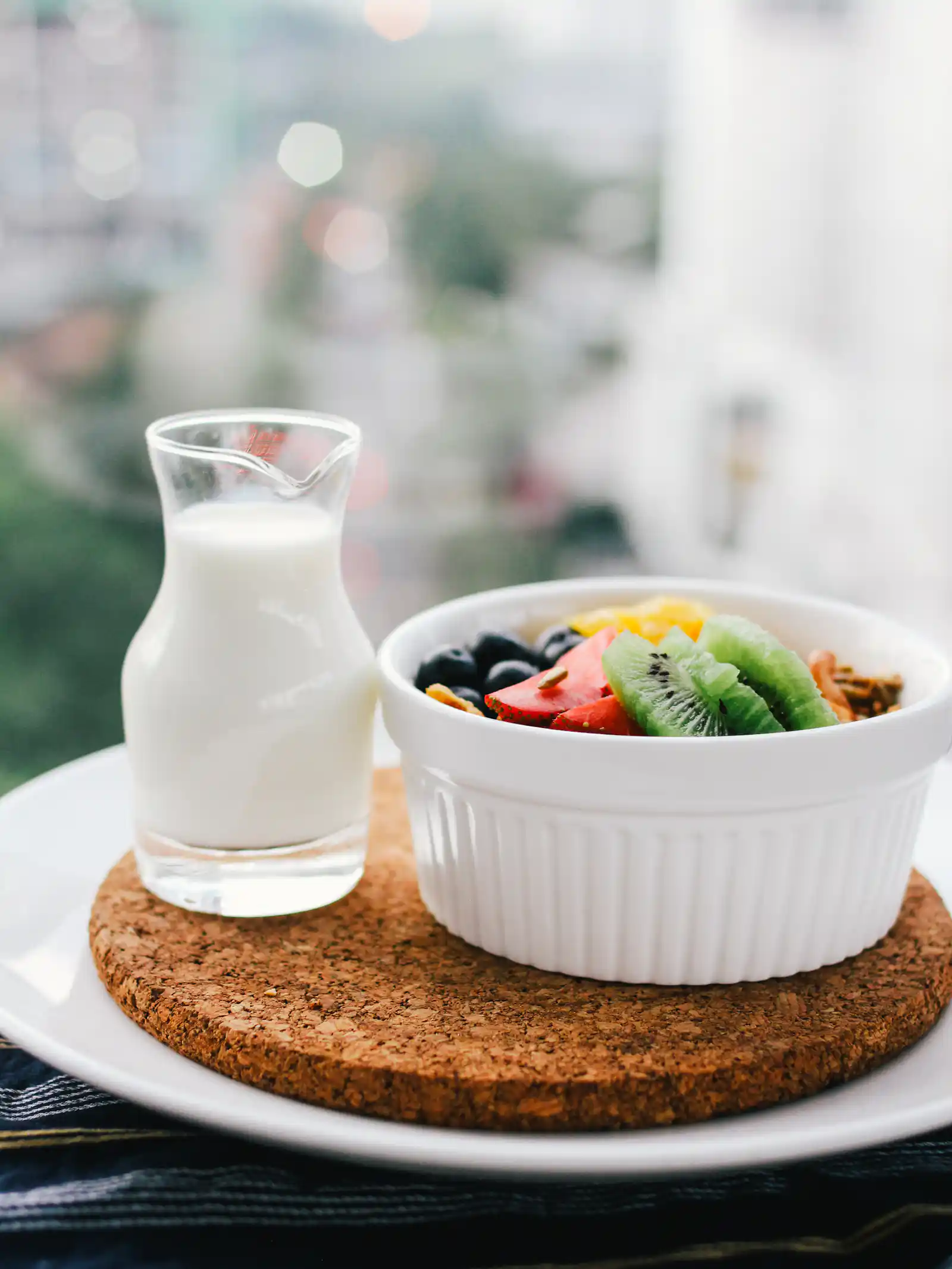 A nutritious breakfast featuring fresh fruits in a white bowl alongside a glass of milk, served on a cork mat with a blurred urban background