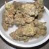 Von Big Buddha Seeds Cherry Moon Pie.Close-up of dried cannabis buds in a white container. The buds are dense and covered with crystalline trichomes, displaying shades of green, orange, and purple.
