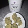 A container of Peace Naturals medicinal cannabis labeled "30:1 GMO Cookies" next to a white lid with several buds of dried cannabis displayed on it.